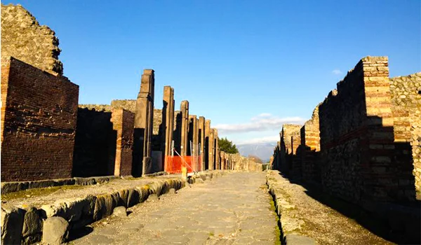 Rua de pedra entre antigas construções e colunas das ruínas arqueológicas de Pompeia, sob um céu azul.