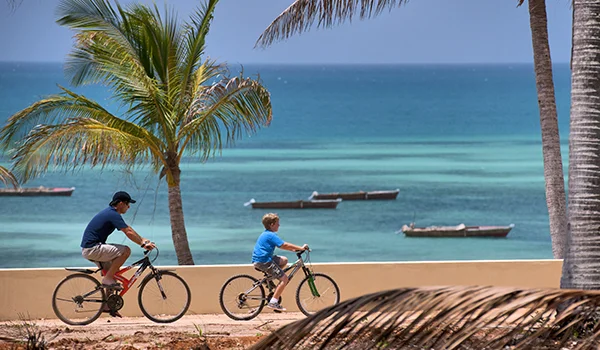 Homem e criança andam de bicicleta à beira-mar, sob palmeiras, com pequenas embarcações sobre águas azul-turquesa ao fundo.