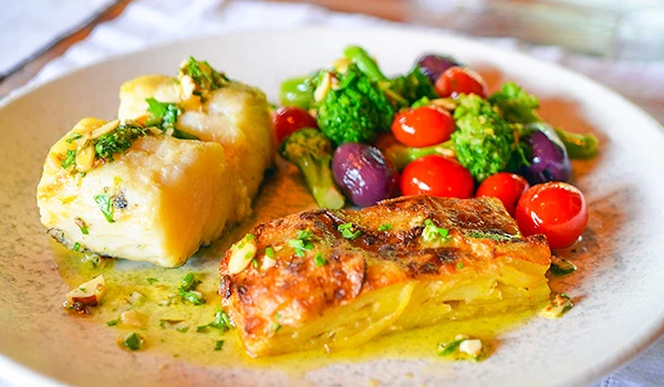 Prato de culinária brasileira com peixe assado, batata ao alho, e salada colorida de tomates, brócolis e uvas vermelhas, servido em um prato rústico. Lua de Mel em Santo Antônio do Pinhal.