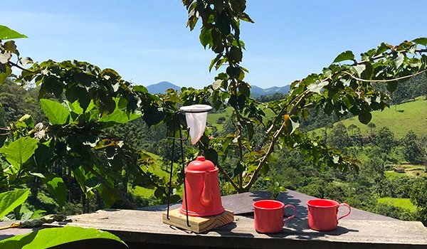 Café feito na estrada com uma cafeteira e duas canecas vermelhas em uma paisagem rural com montanhas ao fundo. Lua de Mel em Santo Antônio do Pinhal.
