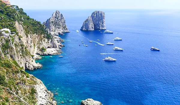 Costa da ilha de Capri, na Itália, com falésias rochosas e vegetação, o mar azul-turquesa e vários barcos próximos aos famosos Faraglioni.