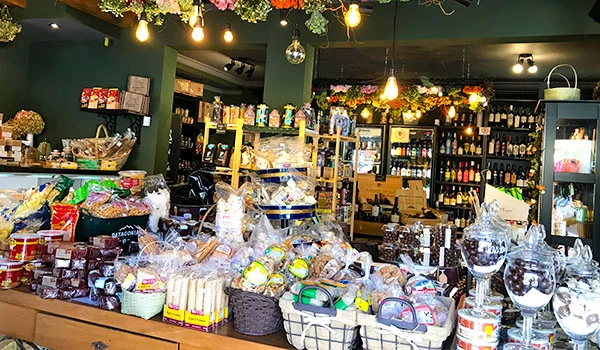Imagem de uma loja de doces e produtos gourmet decorada com luzes e flores, exibindo uma variedade de chocolates, balas, e itens de confeitaria em prateleiras e mesas. Ambiente colorido e acolhedor, ideal para presentes e celebrações.