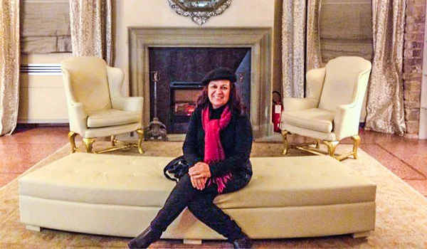 Deusa Rodrigues em um sofá diante de uma lareira ornamentada, em um ambiente interno elegante.