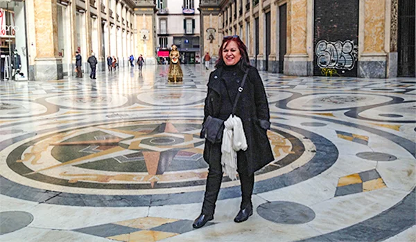 Deusa Rodrigues em pé no interior de uma galeria histórica, cercada por edifícios com colunas e fachadas ornamentadas, sobre um piso de mosaicos com desenhos geométricos e coloridos.