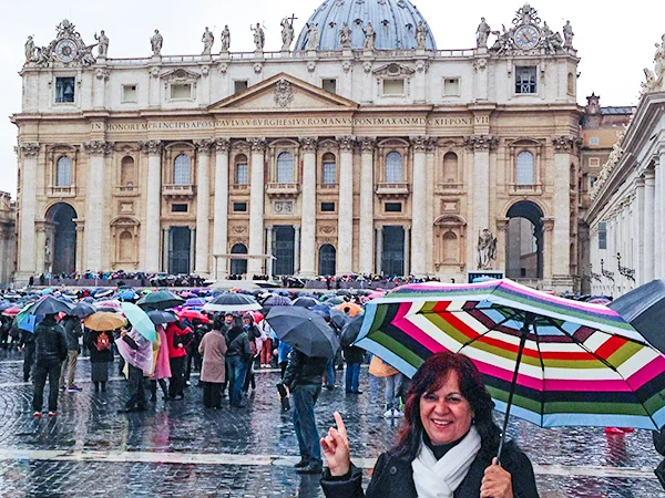 Deusa Rodrigues e segurando um guarda-chuva colorido em frente à Basílica de São Pedro, no Vaticano, enquanto turistas caminham pela praça em um dia chuvoso.