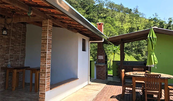 Imagem de uma área externa de uma casa rústica com piso de tijolos, mesa de jantar com cadeira, guarda-sol verde, cercada por natureza verde, ideal para lazer ao ar livre.