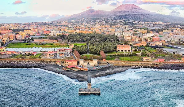 Vista aérea de Nápoles, na Itália, com a costa e o mar em primeiro plano, uma área urbana e arborizada ao centro e o Monte Vesúvio ao fundo.