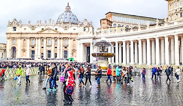 Praça de São Pedro, no Vaticano, com a Basílica de São Pedro ao fundo, colunatas laterais e muitos visitantes caminhando pelo local.
