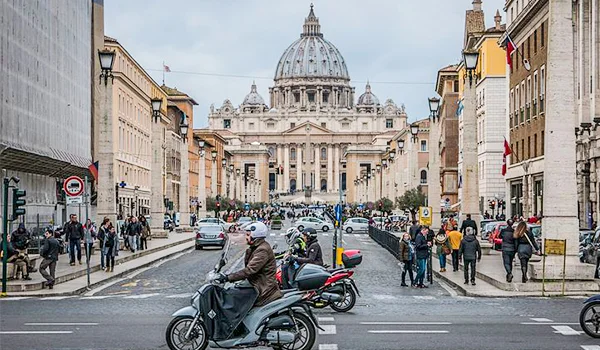 Vista de uma avenida de Roma com a Basílica de São Pedro ao fundo, destacando sua grande cúpula. Pessoas caminham pelas calçadas enquanto motocicletas, carros e ônibus circulam pela rua. Lua de Mel na Itália.