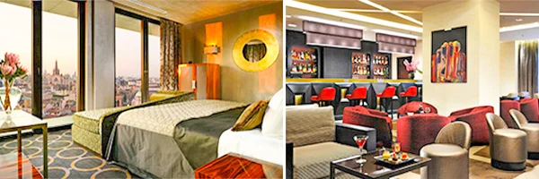 Quarto de hotel moderno com cama de casal e grandes janelas com vista para a cidade. Ao lado, lounge elegante com poltronas, sofás, mesas, bar e obras de arte nas paredes.
