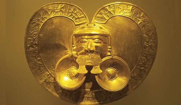 Arte antiga de uma peça de ouro com coração, escrita mystica ao redor e uma face estilizada ao centro com dois tambores, símbolo de cultura e espiritualidade. Lua de Mel na Colômbia.