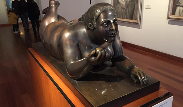 Escultura de uma mulher deitada com expressão tranquila, feita de bronze, exibida em um museu de arte, com molduras de quadros ao fundo. Lua de Mel na Colômbia.