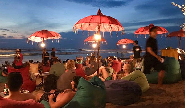 Pessoas curtindo um evento na praia ao entardecer, com guarda-chuvas decorativos vermelhos e público sentado em almofadas, criando uma atmosfera relaxante e animada para eventos na praia ao pôr do sol em Bali.