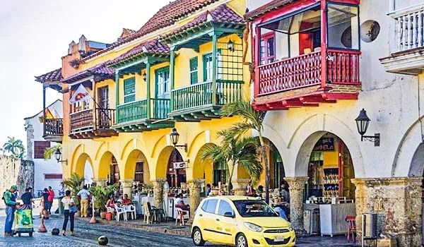 Fachada colorida de edifícios históricos em uma rua movimentada de Cartagena, Colômbia, com turistas apreciando a arquitetura vibrante e restaurantes ao ar livre.