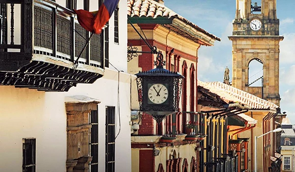 Imagem de uma rua charmosa com arquitetura colonial, featuring relógios de rua em fachadas coloridas e um relógio na torre ao fundo, típicas de uma cidade histórica.