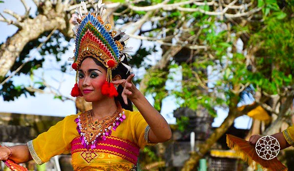 Mulher vestida com traje tradicional e adereço de cabeça colorido, participando de festa cultural ao ar livre, com árvores ao fundo.