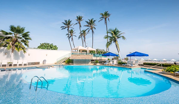 Piscina de resort com águas azuis, rodeada por cadeiras de descanso, guarda-sóis azuis e palm trees ao fundo, sob um céu claro, ideal para lazer e férias em clima tropical.