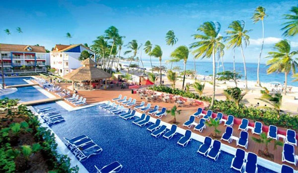 Piscina de resort com vista para a praia caribenha, palmeiras ao redor, cadeiras de praia brancas, azul do mar e céu claro ao fundo.