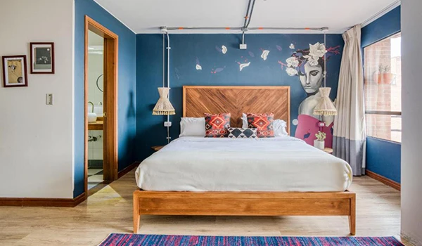 Quarto de hotel moderno com decoração artística na parede azul, cama de madeira ampla e detalhes de arte colorida, ambiente acolhedor e bem iluminado. Lua de Mel na Colômbia.