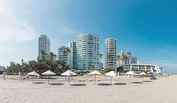 Imagem de praia com areia branca, espreguiçadeiras e guarda-sóis, ao fundo, arranha-céus modernos e palmeiras, sob céu claro e ensolarado. Ideal para turismo e lazer na orla.