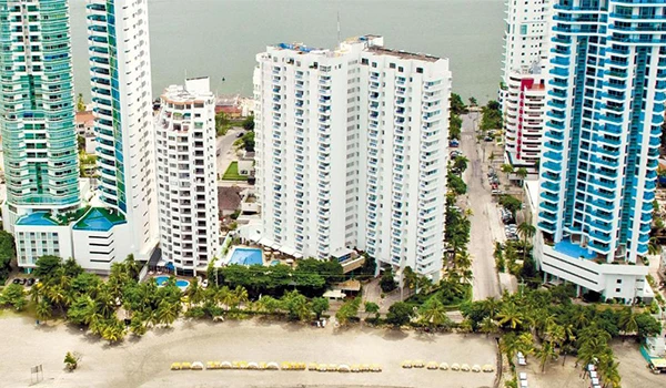 Imagem de prédios residenciais na orla de uma cidade costeira, com vista para o mar ao fundo e áreas verdes entre as construções.