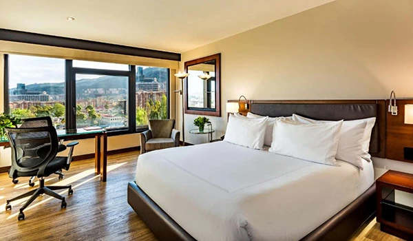 Quarto de hotel moderno com vista para a cidade, cama confortável, cadeira de escritório e janela grande, ideal para hospedagem de qualidade. Lua de Mel na Colômbia.
