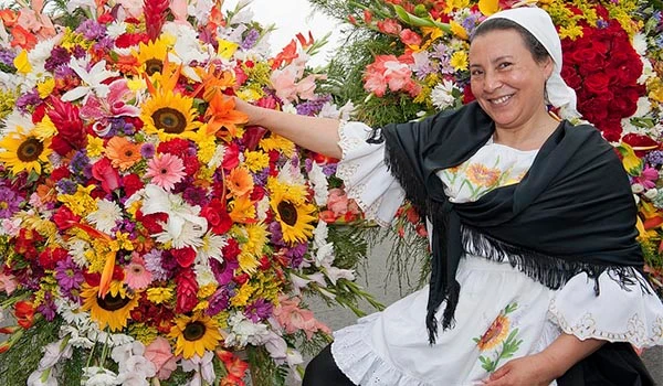 Mulher sorridente vestido tradicional e lenço preto rodeada de um grande arranjo de flores coloridas, incluindo girassóis, rosas e cravos, em celebração de eventos culturais ou religiosos.