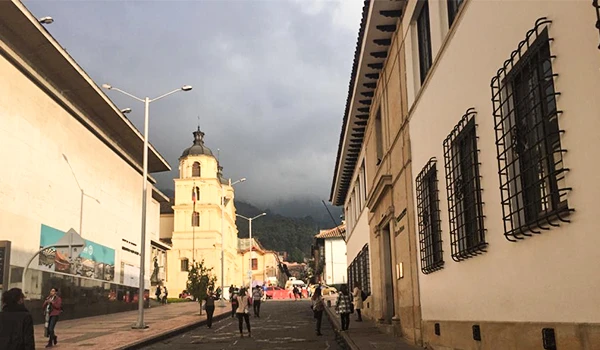 Cenário urbano em uma rua histórica com uma igreja ao fundo e pessoas caminhando ao longo do dia, em uma cidade com relevo montanhoso ao fundo. Lua de Mel na Colômbia.