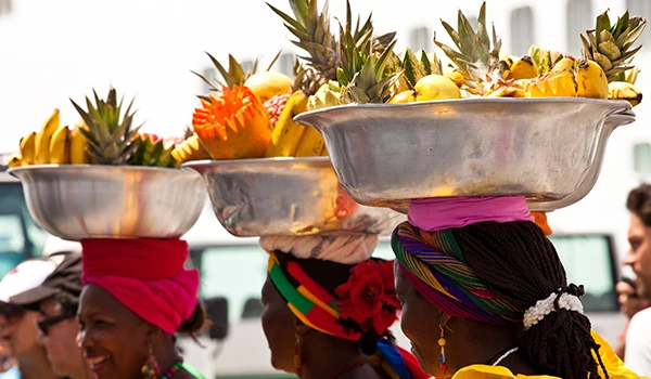 Mulheres colombianos carregando cestos cheios de frutas tropicais, incluindo abacaxi, em um mercado ao ar livre, vestindo roupas coloridas e tecidos tradicionais.