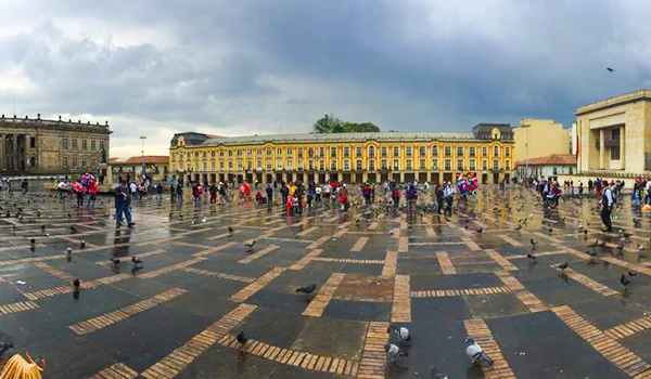 Imagem da Praça Principal de Bogotá, Colômbia, com visitantes, pavimentação de pedra, prédios históricos ao fundo e céu nublado. Uma vista destacada de um espaço turístico e cultural. Lua de Mel na Colômbia.