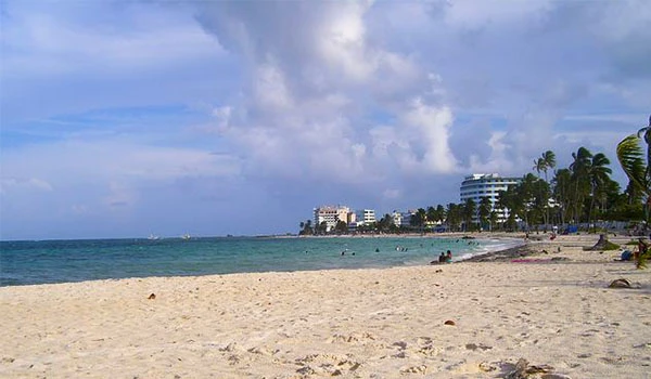 Praia de areia branca e mar azul com céu parcialmente nublado, vista panorâmica do litoral de uma cidade costeira com edifícios e palmeiras ao fundo