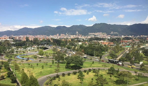 Vista panorâmica de um parque urbano em uma cidade brasileira, com áreas verdes, árvores, vias de trânsito intenso e uma cadeia de montanhas ao fundo, sob céu parcialmente nublado. Lua de Mel na Colômbia.