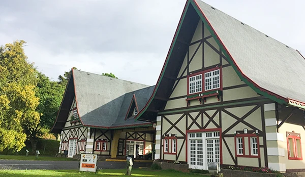 Imagem de uma casa de estilo colonial com detalhes em madeira, janelas brancas e telhado de duas águas, rodeada por árvores verdes. 