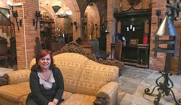 Imagem de Deusa Rodrigues sentado em um sofá antigo de tecido dourado em um ambiente que lembra uma sala de eventos ou restaurante com decoração rústica, paredes de tijolos expostos, luminárias pendentes e móveis tradicionais. Harry Potter em Campos do Jordão.