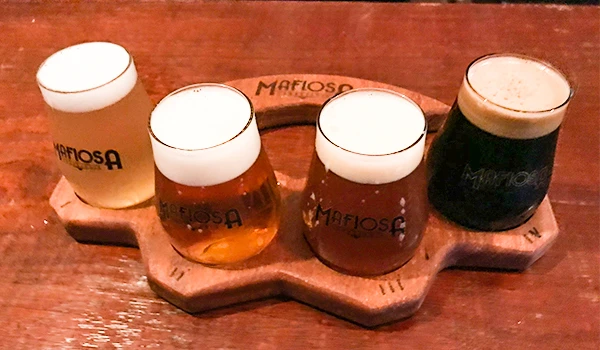 Um conjunto de quatro copos de cerveja artesanal da marca Mafiosa, apresentando diferentes tipos de cervejas em um suporte de madeira.