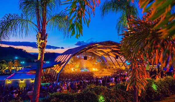 Uma vista deslumbrante de um festival de música ao ar livre, com um grande público sob um céu azul e palmeiras iluminadas, criando um ambiente vibrante e festivo. Balada Laroc em Valinhos.