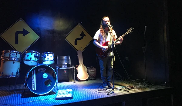 Músico se apresentando em um palco com bateria e guitarra, cercado por placas de sinalização. Ambiente escuro e iluminação de palco.