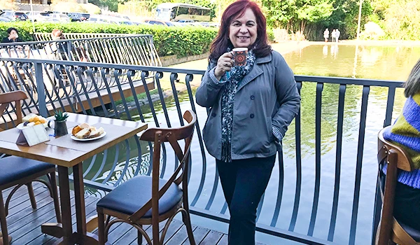 Deusa Rodrigues segurando uma caneca em um ambiente ao ar livre, com mesas e natureza ao fundo. Café colonial no castelo em vinhedo.