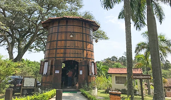 Adega Santa Cecília imagem de uma charmosa construção em formato de barril, localizada em um ambiente natural, com árvores e plantas ao redor. O lugar possui uma entrada com pessoas e estrutura típica de um restaurante ou ponto turístico.