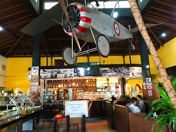 Ambiente de um restaurante com decoração temática de aviação, destacando um modelo de avião suspenso no teto, bar e área de refeições.