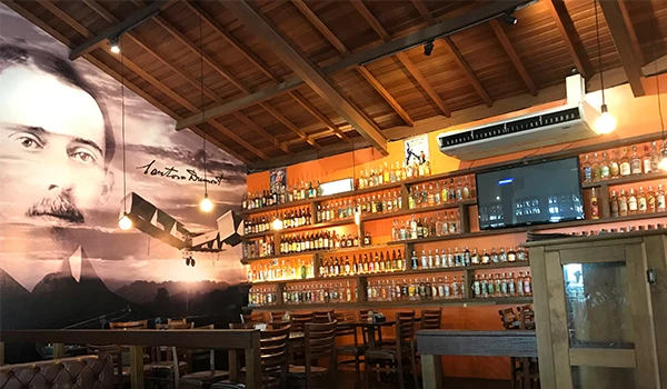 Interior de restaurante com decoração que homenageia Santos Dumont, incluindo uma parede com sua imagem e prateleiras cheias de garrafas. O ambiente é acolhedor e rústico.