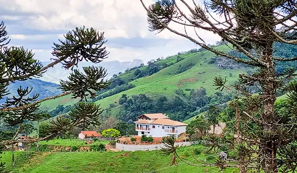 Paisagem de uma fazenda rodeada por árvores e colinas verdes, com uma casa branca ao centro e céu parcialmente nublado ao fundo em zona rural.