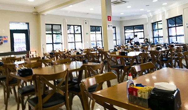 Imagem de um restaurante vazio, com mesas de madeira organizadas, amplas janelas proporcionando luz natural e um ambiente acolhedor.
