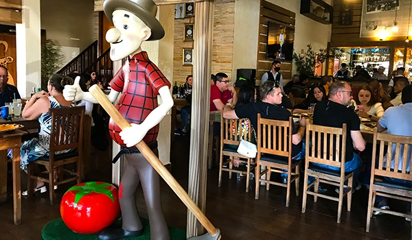 Estátua de um personagem caipira, sorridente, segurando uma pá e com um tomate em destaque, em ambiente de restaurante cheio de pessoas.