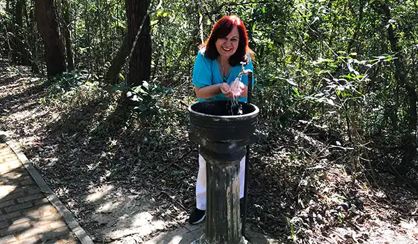 Deusa Rodrigues enquanto enche a mão com água em uma fonte natural no meio da natureza, rodeada por árvores e folhas secas.