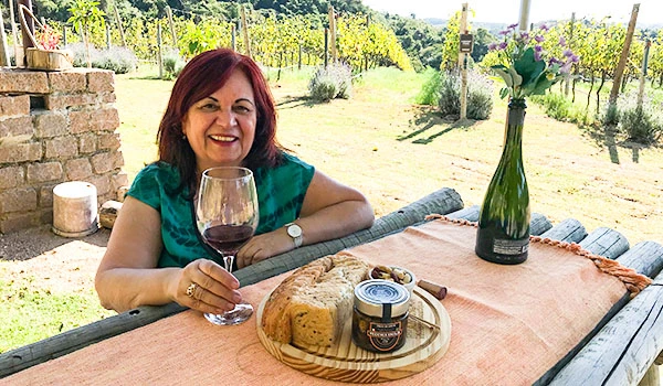 Deusa Rodrigues com taça de vinho tinto, pão e petiscos em mesa ao ar livre, rodeada por vinhedos e natureza.