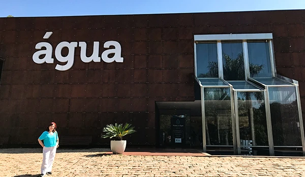 Imagem de uma mulher de blusa azul em frente a um edifício com a palavra 'água' em destaque na fachada. Local ideal para eventos e reflexões sobre água.