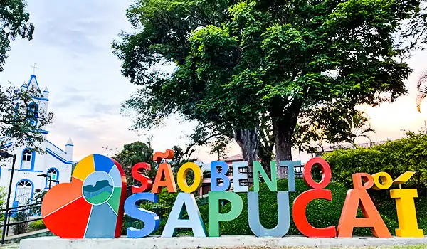 Imagem colorida sobre que fazer em São Bento do Sapucaí, com uma placa de sinalização que destaca o nome da cidade, árvores ao fundo e céu ao entardecer.