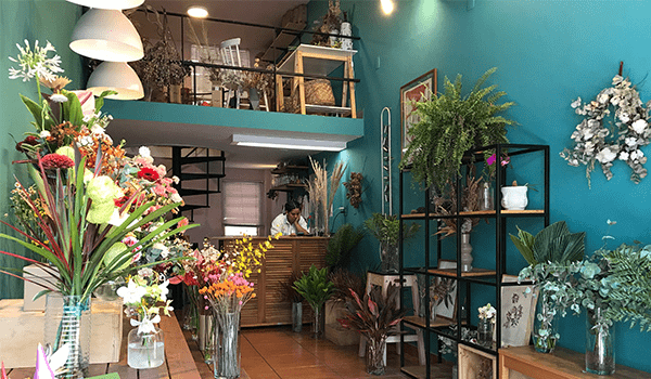 Foto do interior da loja Arranjo Floral, toda na cor verde, com prateleiras e balcão com flores e uma funcionária.