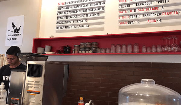 Foto de um balcão com atendente da Cafeteria Gourmet Naïf Café, tendo acima na parede uma placa do cardápio com preços.